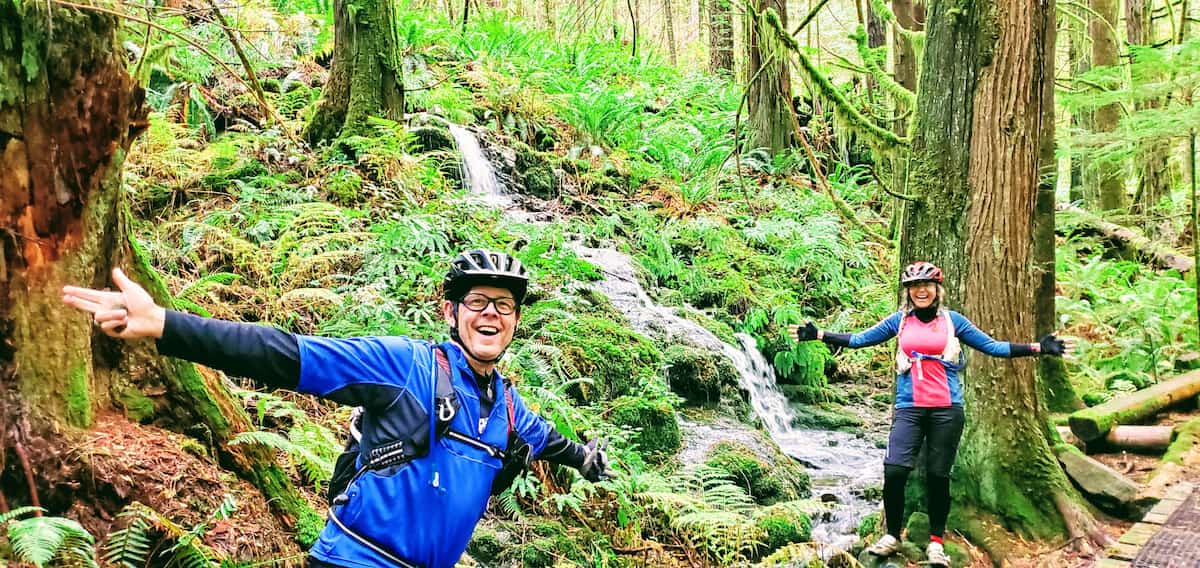 travel bloggers Rob and Mash are standing in their biking gear with a small waterfalls between them in a very green Elk Falls parks which is one of the best things to do on Vancouver Island