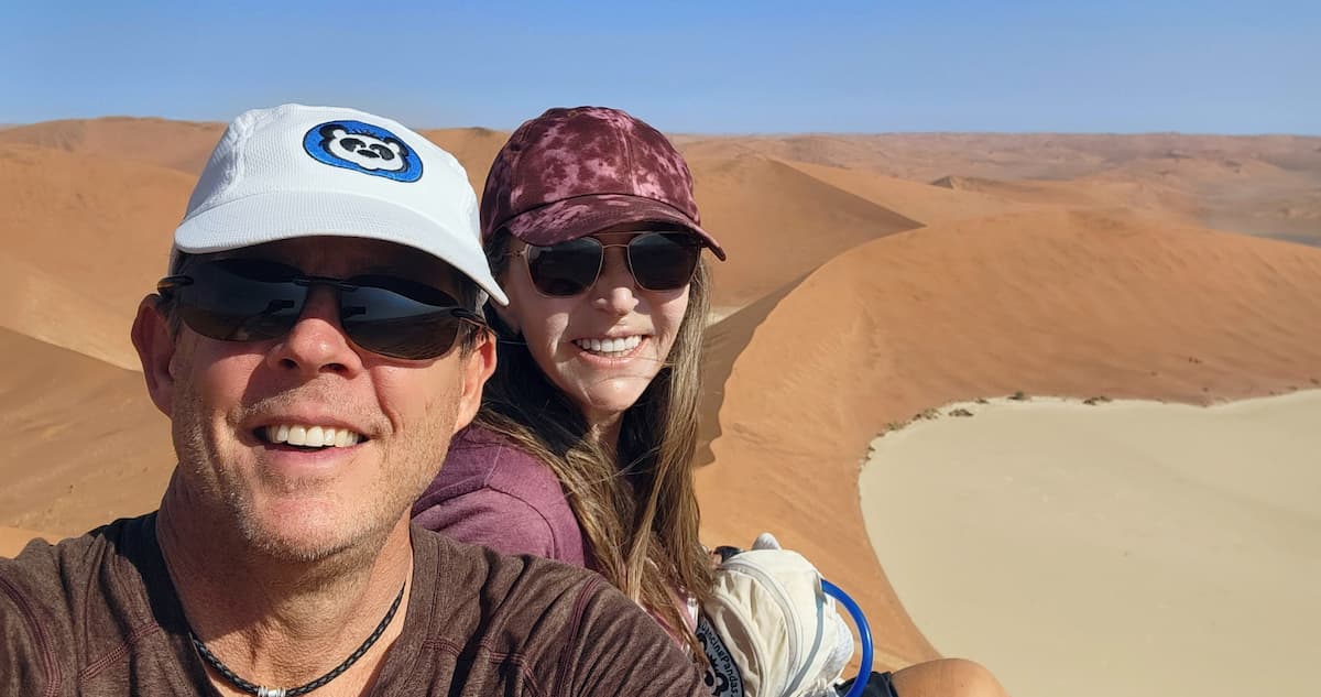 Masha and Rob smiling for a selfie on top of a massive red sand dune overlooking a white clay pan at Sossusvlei, exploring top Namibia tourist attractions.